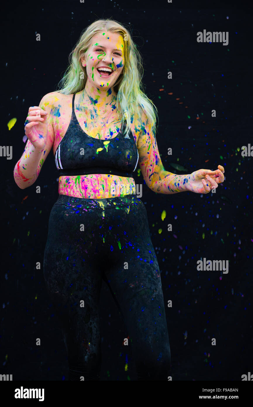 A laughing happy young blonde haired woman girl female  model standing against a black background splattered with splashes of multicoloured paint Stock Photo