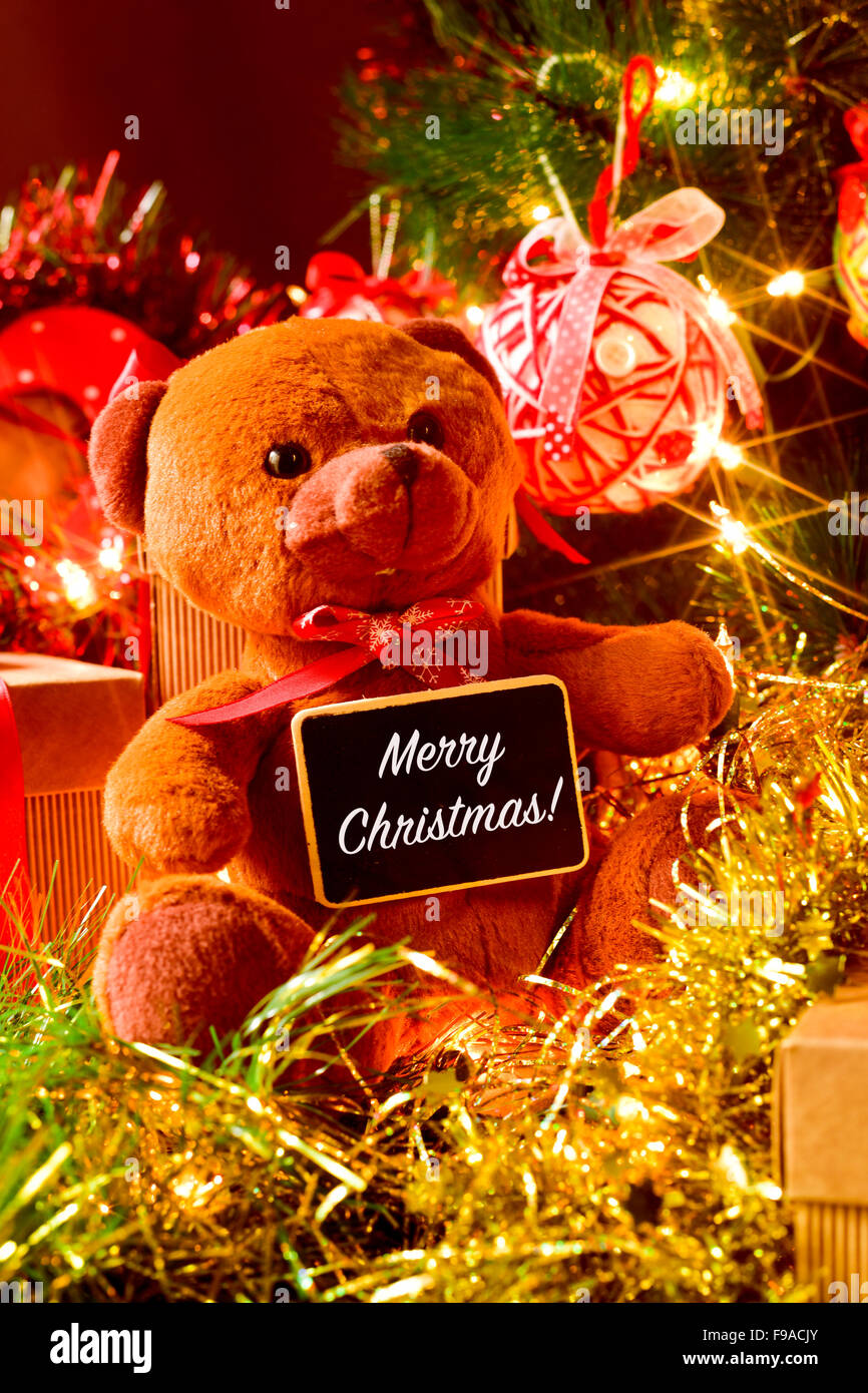 closeup of a brown teddy bear with a chalkboard with the text merry christmas, and some gifts under a christmas tree ornamented Stock Photo