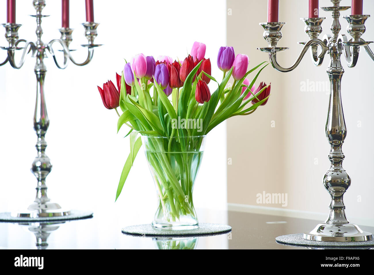 A clear glass vase of red, move and pink tulips on a glass table with two silver candlesticks Stock Photo