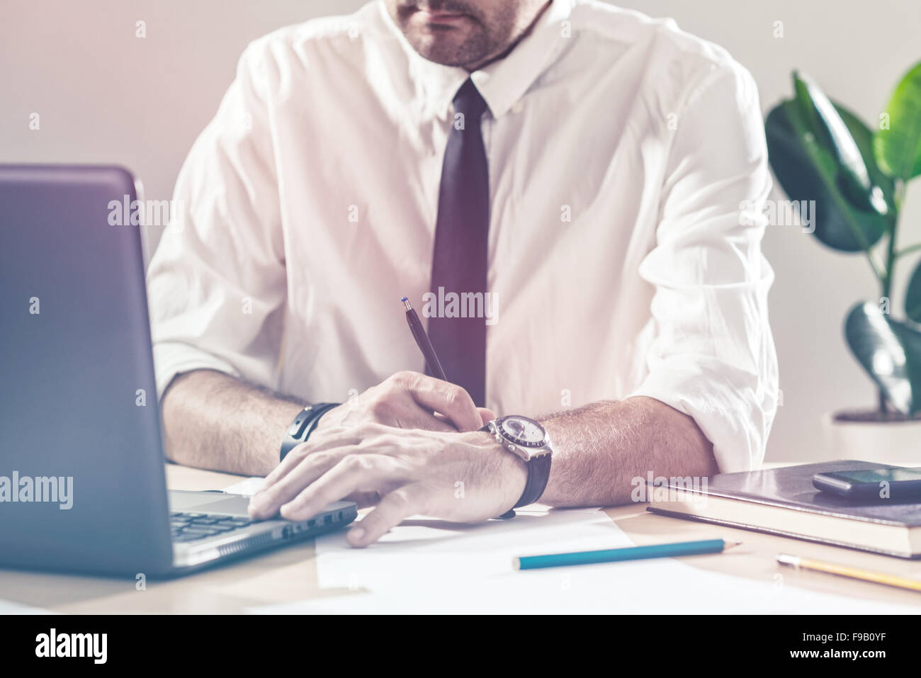 Businessman writing notes and using laptop, working overtime at office desk, multitasking and project deadline concept, retro to Stock Photo