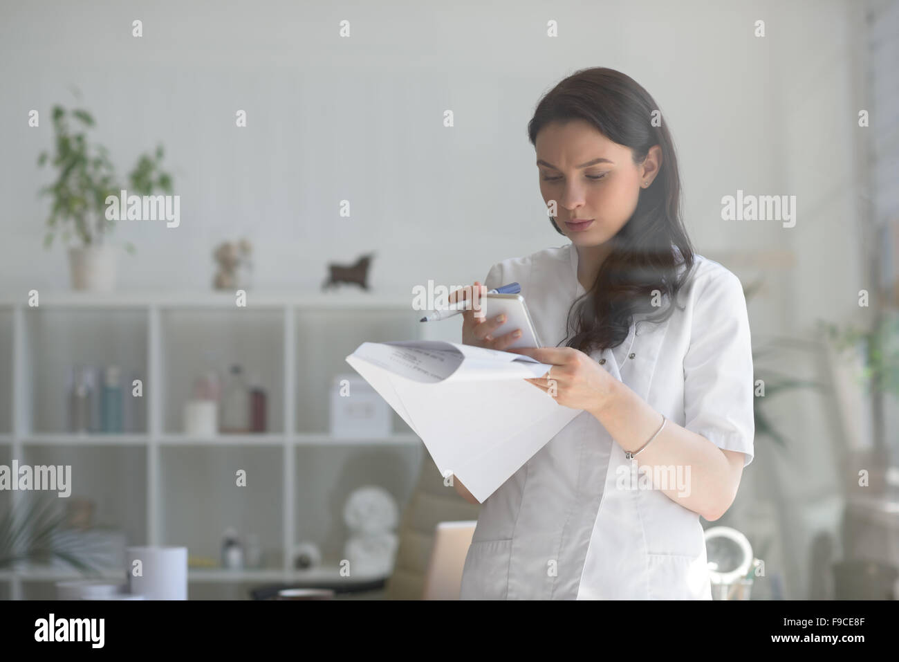 Doctor at her medical office using mobile phone and holding papers. View through the glass Stock Photo