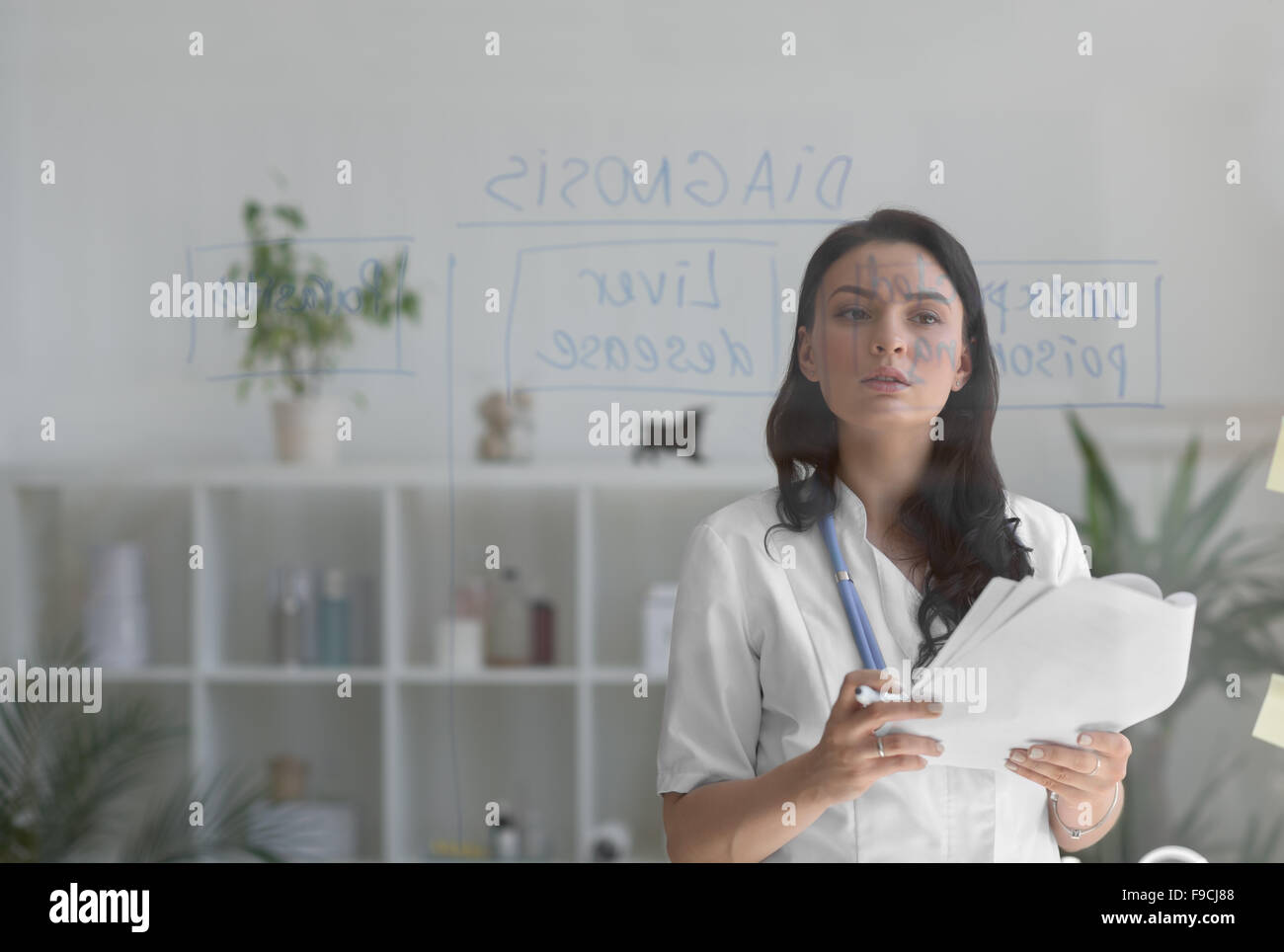Female medical doctor working at clinic office. Writing on glass whiteboard symptoms and test results of her patient to diagnose Stock Photo