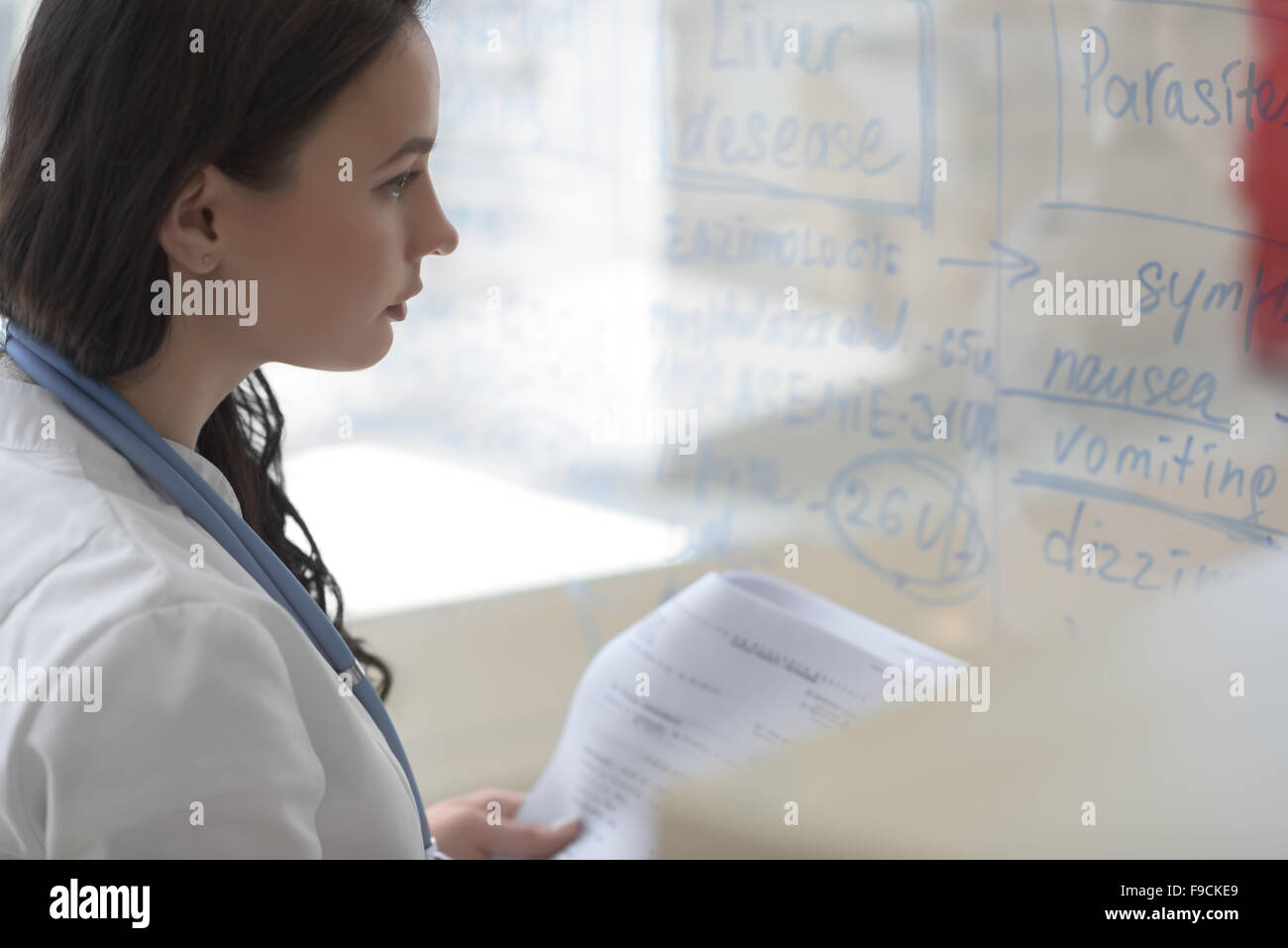 Female medical doctor working at clinic office. Writing on glass whiteboard symptoms and test results of her patient to diagnose Stock Photo