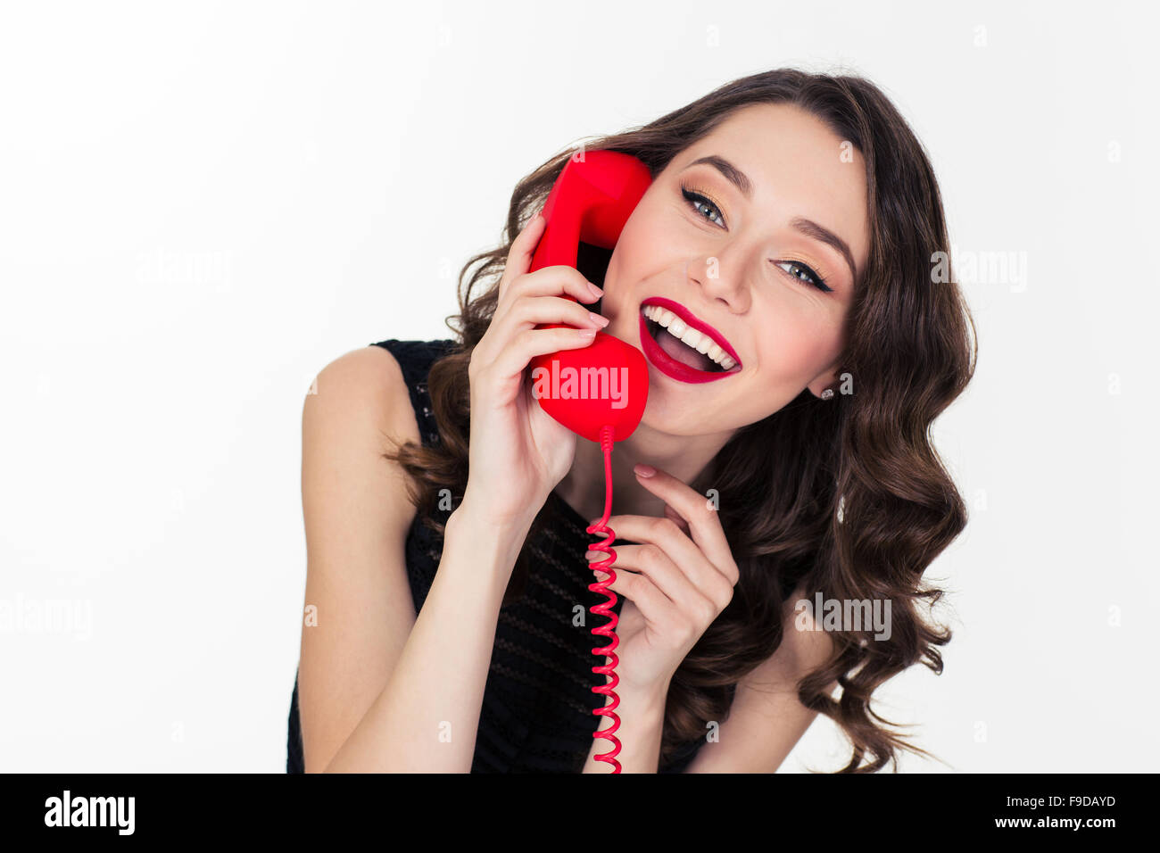 Cheerful attractive curly retro styled young woman talking on red telephone isolated over white background Stock Photo