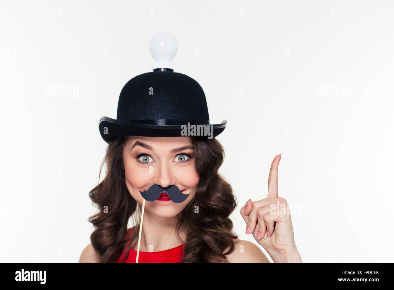 Happy comical young curly girl in amusing black hat with light bulb pointing up and using fake moustache props Stock Photo