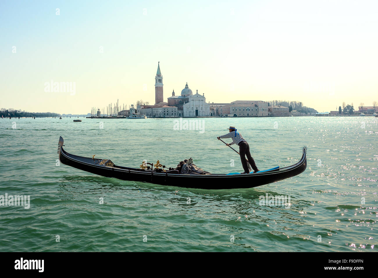 Gondolier in a gondola rides on Canal Grande in a hat with a blue ribbon and a typical striped singlet, Basilica in the backgrou Stock Photo