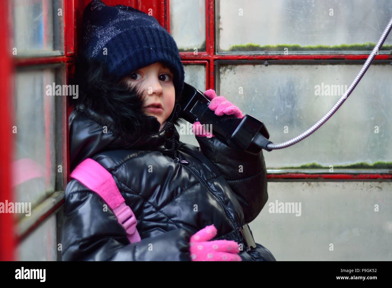 Child in a red telephone box, on the phone. A girl pretends to make a call in a public phone box, dressed warmly for winter Stock Photo