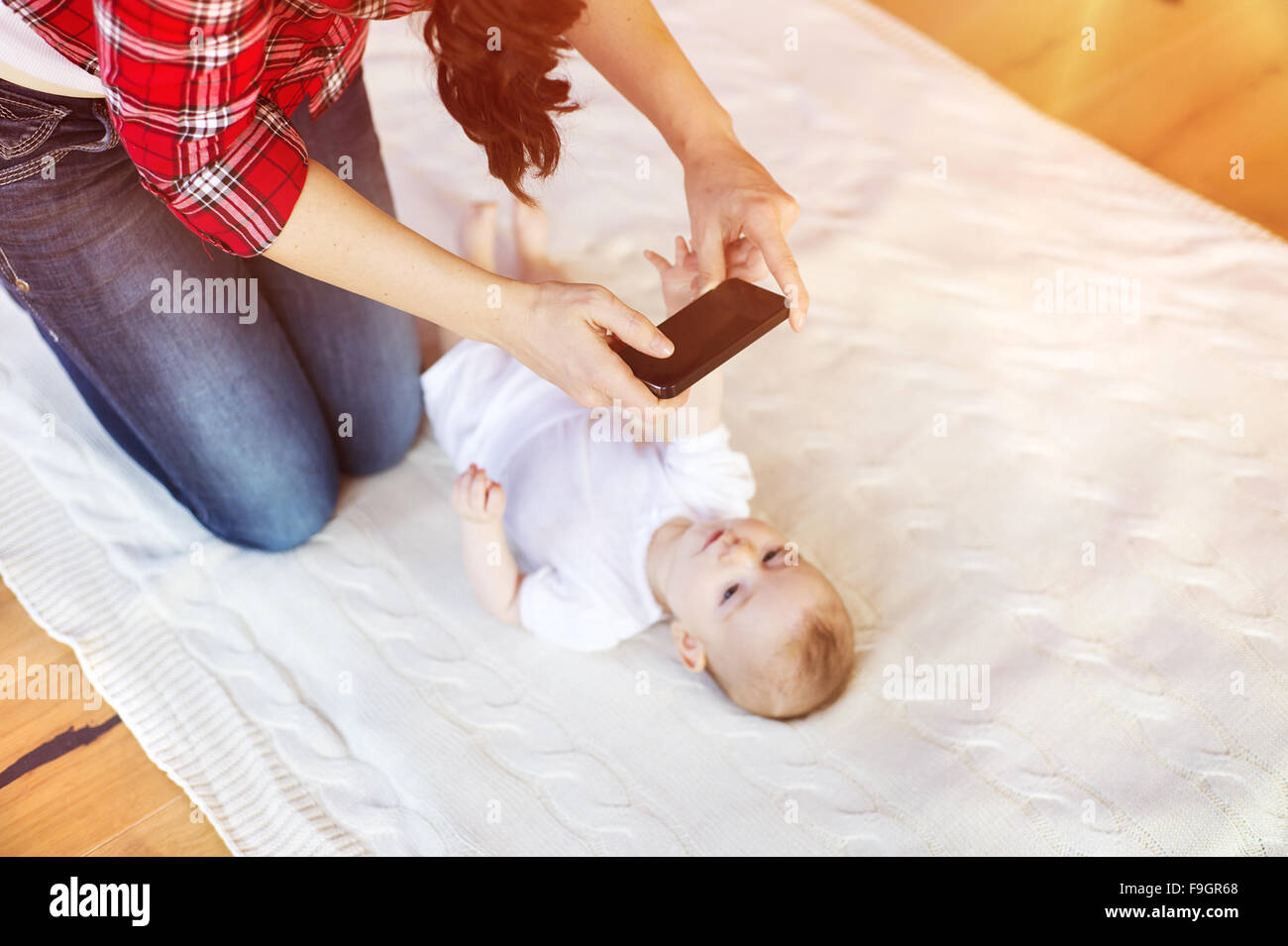 Young mother taking a picture of her cute little baby girl lying on a blanket in a living room. Stock Photo