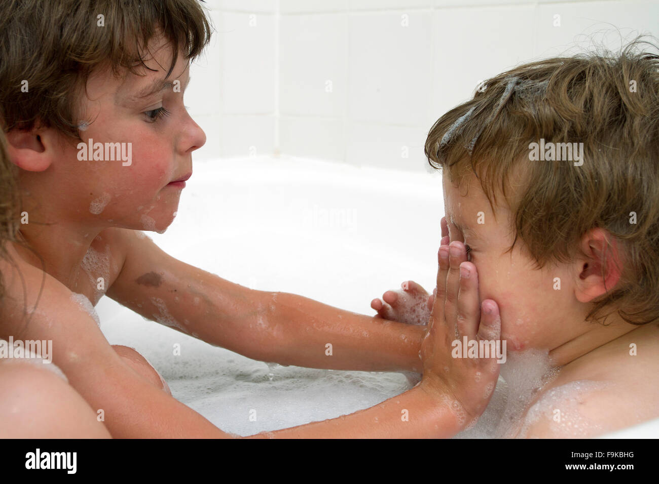 Two toddler boys having fun with bubbles in the bath. Older brother washing his younger sibling's face. Stock Photo
