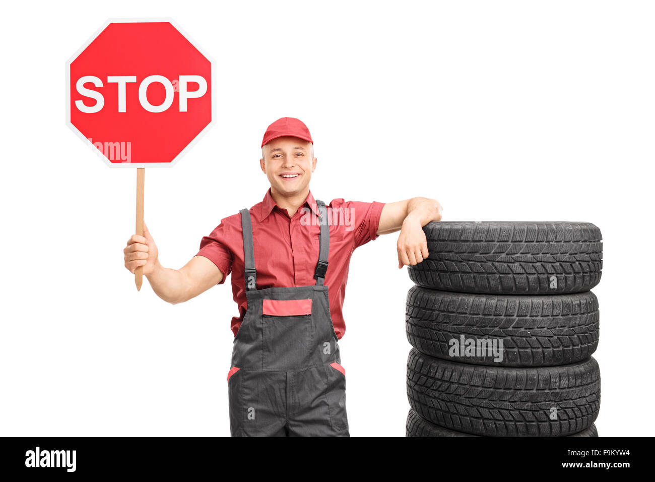 Young male mechanic holding a big red stop sign and leaning on a stack of tires isolated on white background Stock Photo