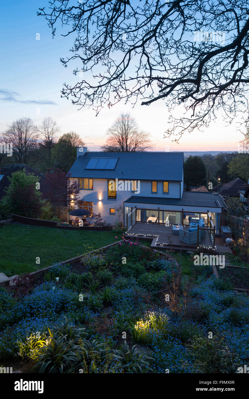 House from the rear at dusk showing the terrace, extension and garden. A modern famly house with large full length glass panels. Lights in the twilight. Solar panels on the roof. Stock Photo