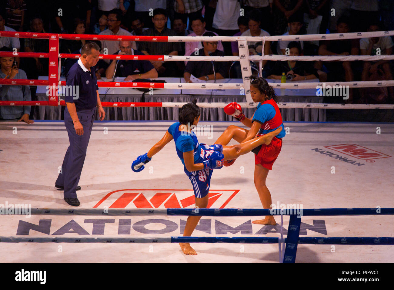 Female muay thai kickboxer in blue kick jab kicking oppenent is blocked by the arm as male referee officiates at amateur outdoor Stock Photo