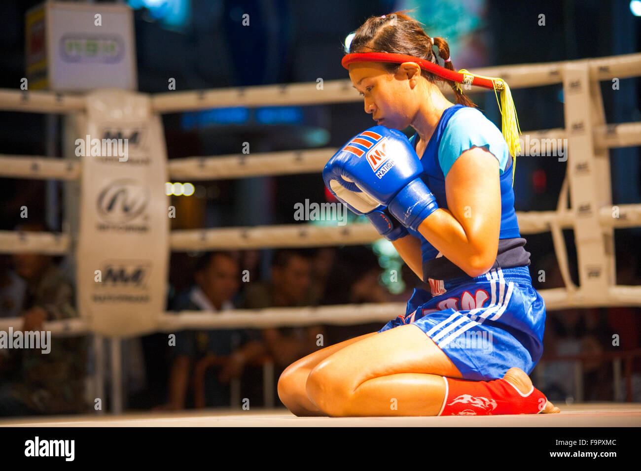 Female muay thai fighter kneeling, gloves raised, reflecting in a kickboxing ritual called the wai khru meant to honor teachers Stock Photo