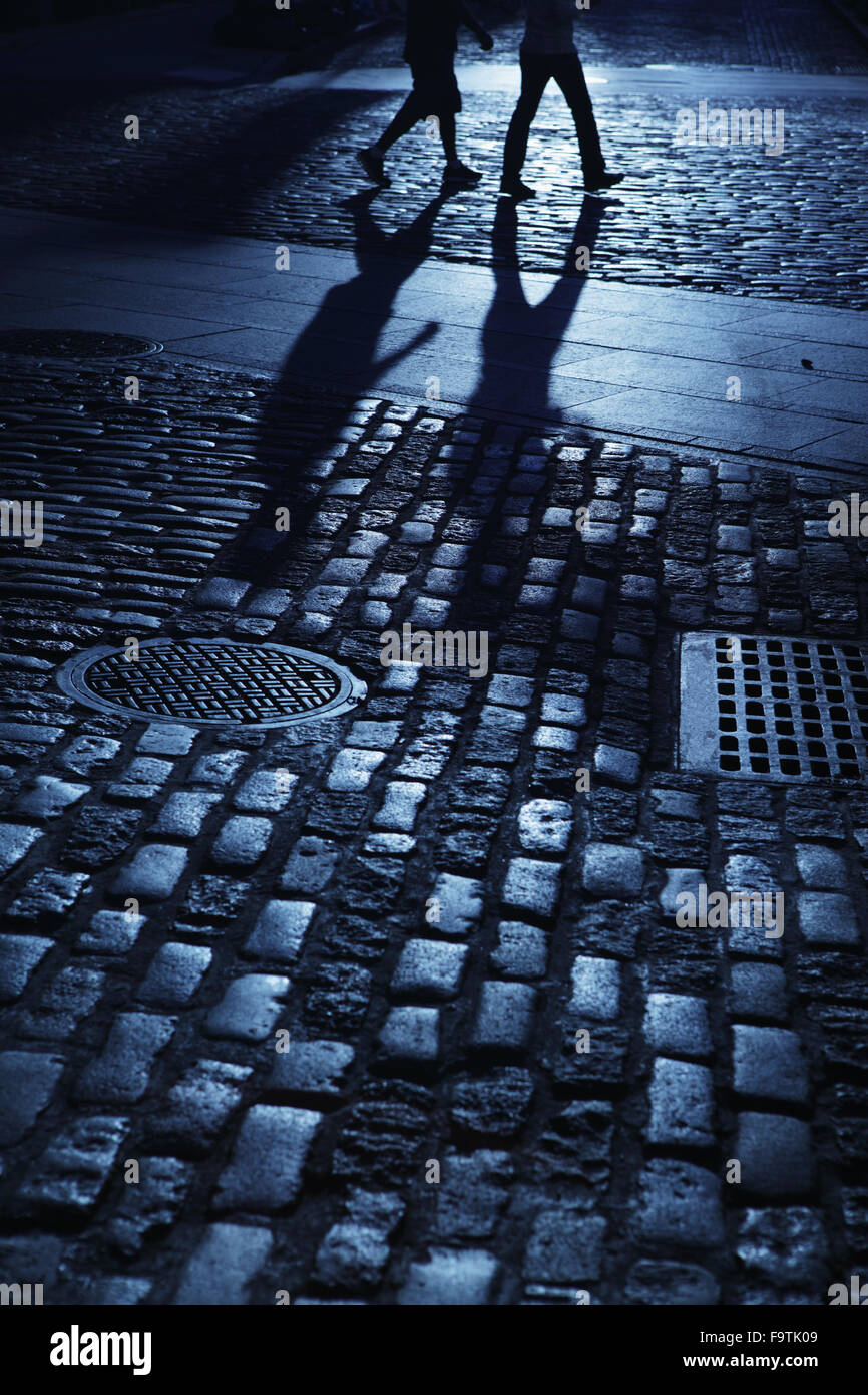 Two young people walking in blue night shadows on cobblestone road Stock Photo