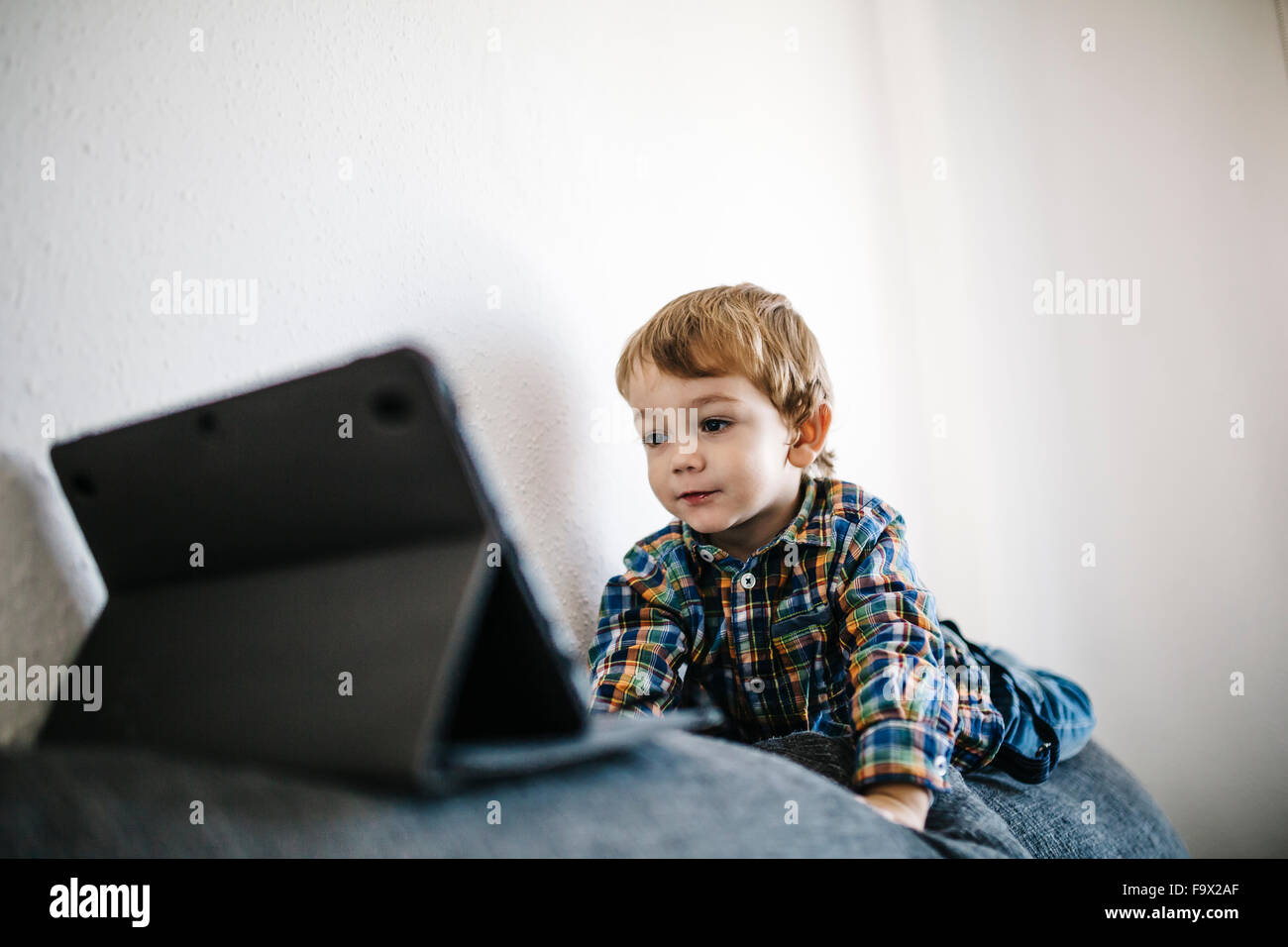 Portrait of little boy lying on backrest of a couch looking at digital ...