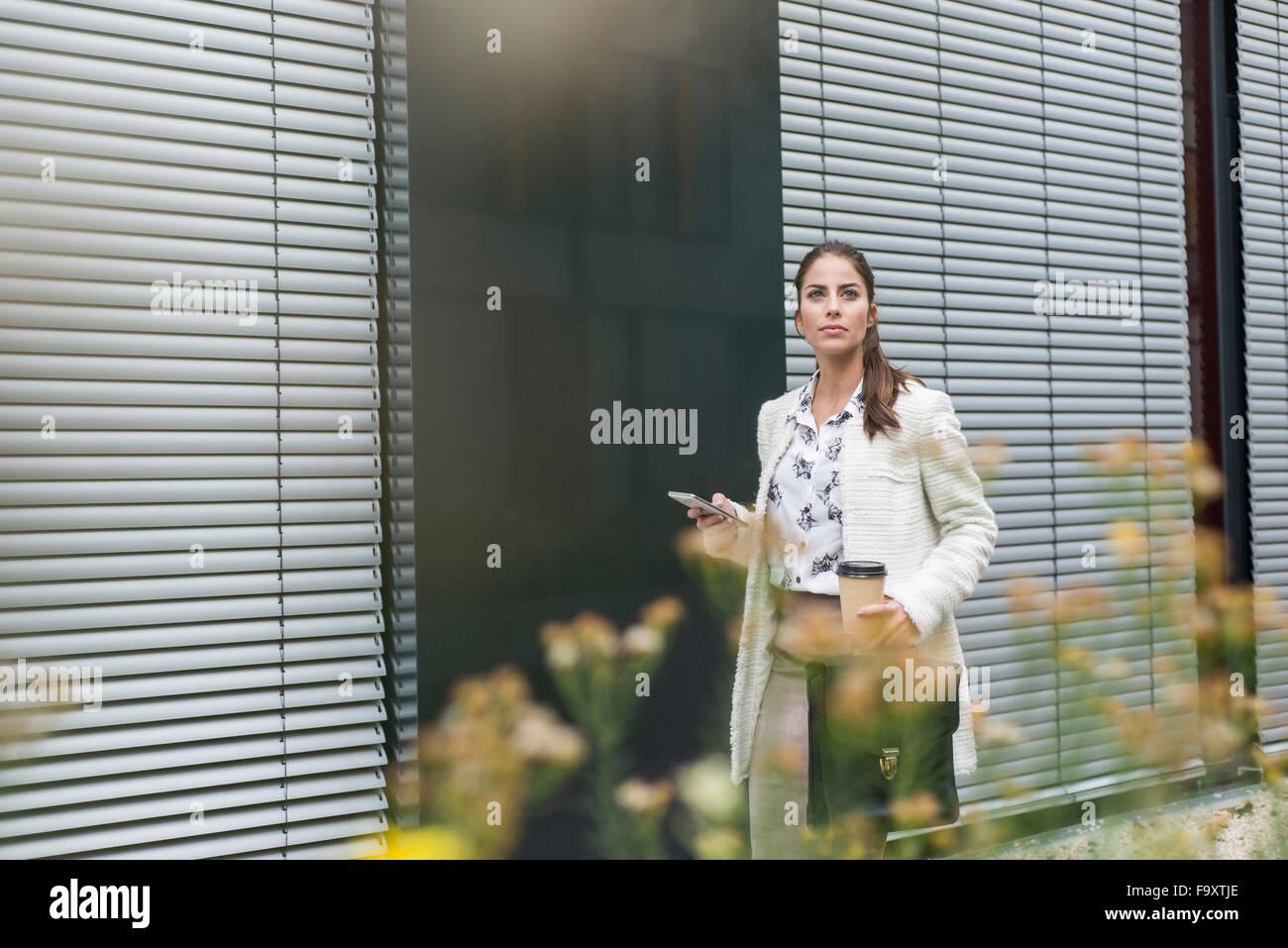 Young businesswoman outdoors holding cell phone and coffee to go Stock Photo
