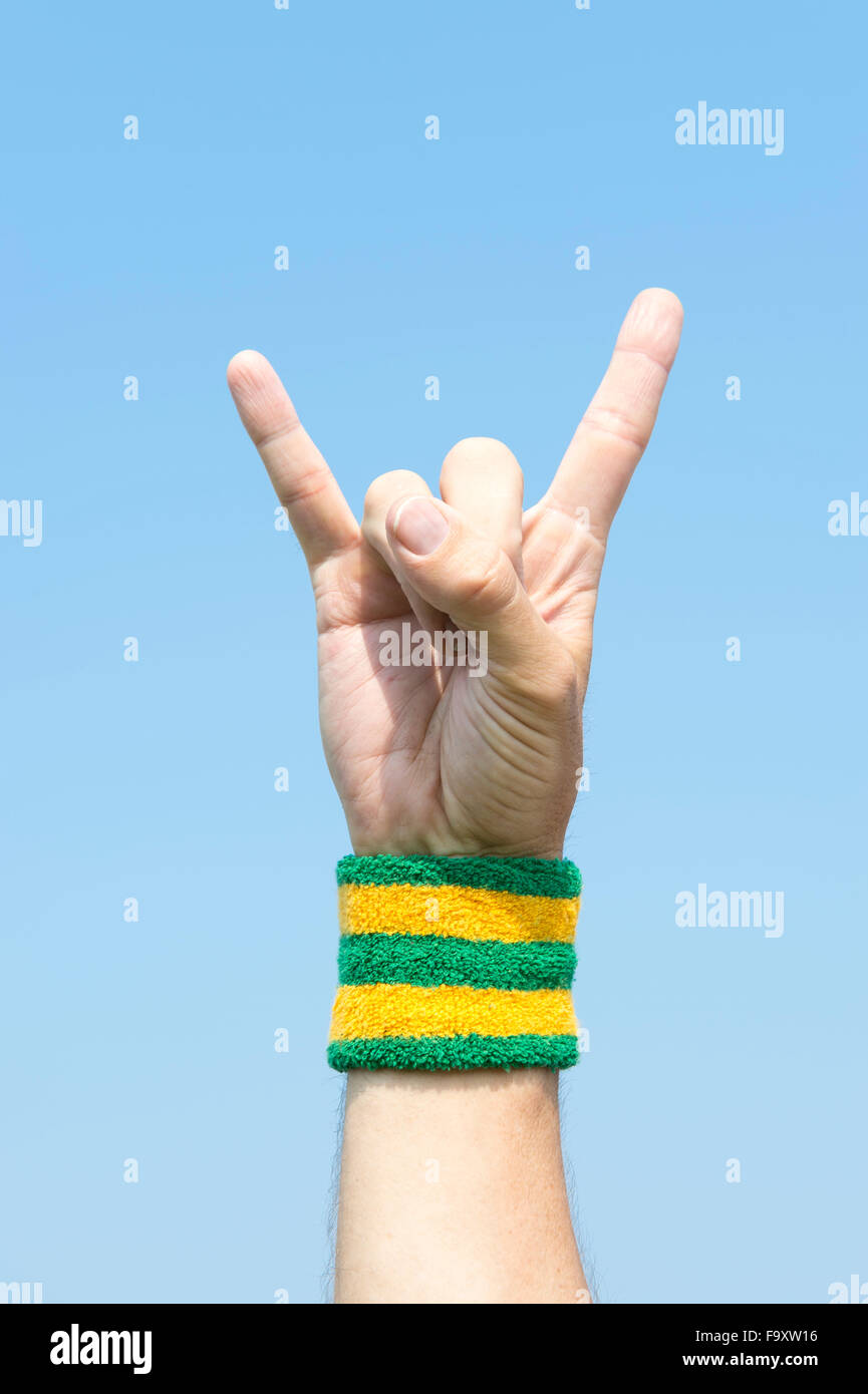 Hand of cool athlete with Brazil colored wristband holding up rocker sign devil horns victory fingers against clear blue sky Stock Photo