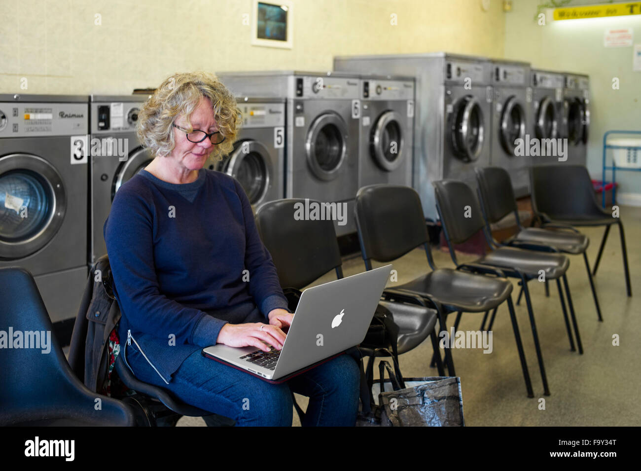 Older woman on Macbook Air in launderette Stock Photo