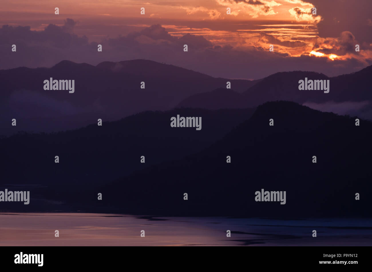 Silhouette of Mountains Above the Water under purple orange cloudscape clouds at sunset Stock Photo