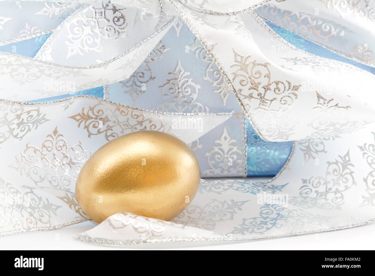 Ice blue wrapped package with silver and white ribbon placed behind gold nest egg. Stock Photo
