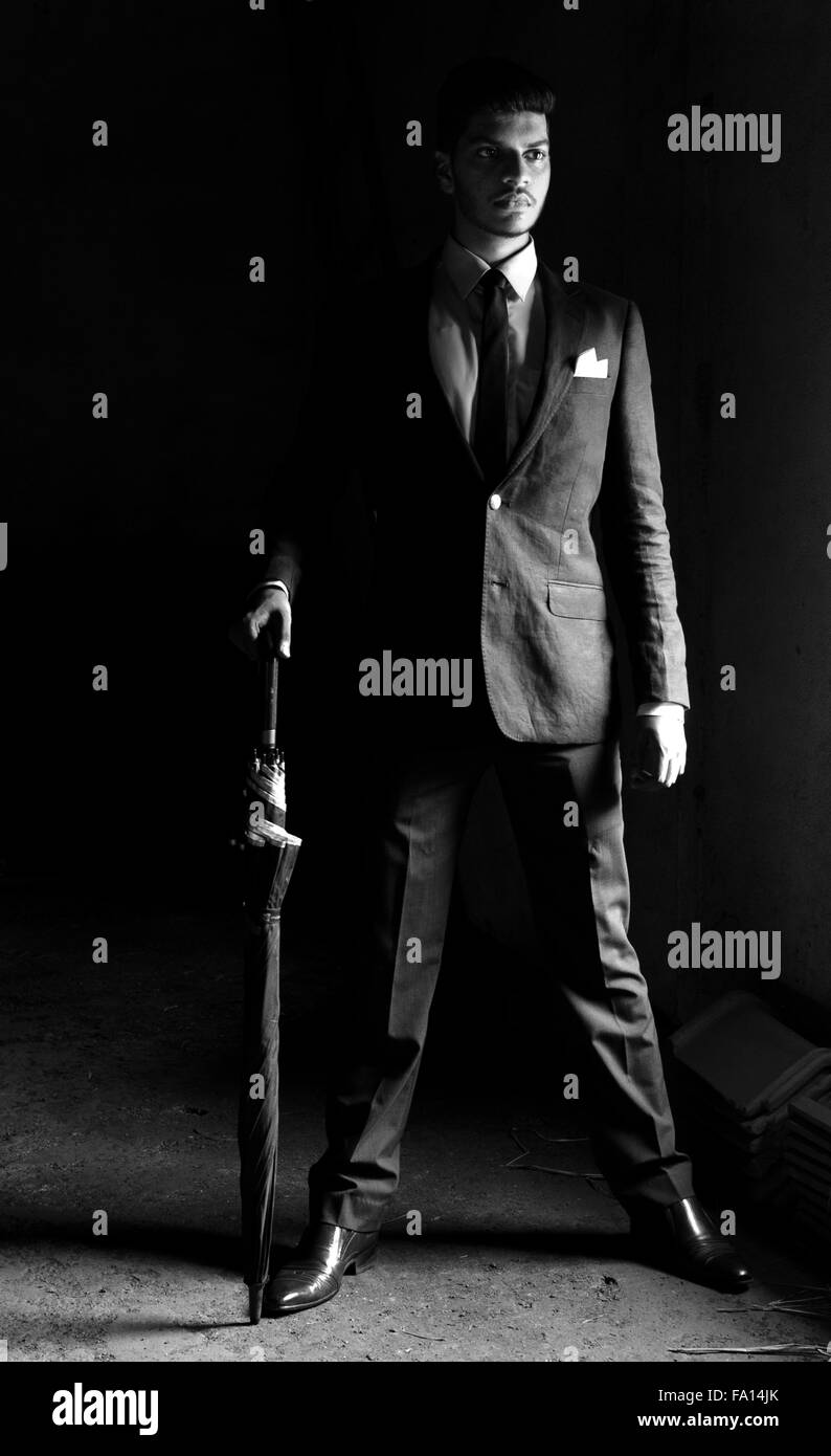 Black and White portrait of a young man dressed in black suit posing with umbrella over dark background Stock Photo