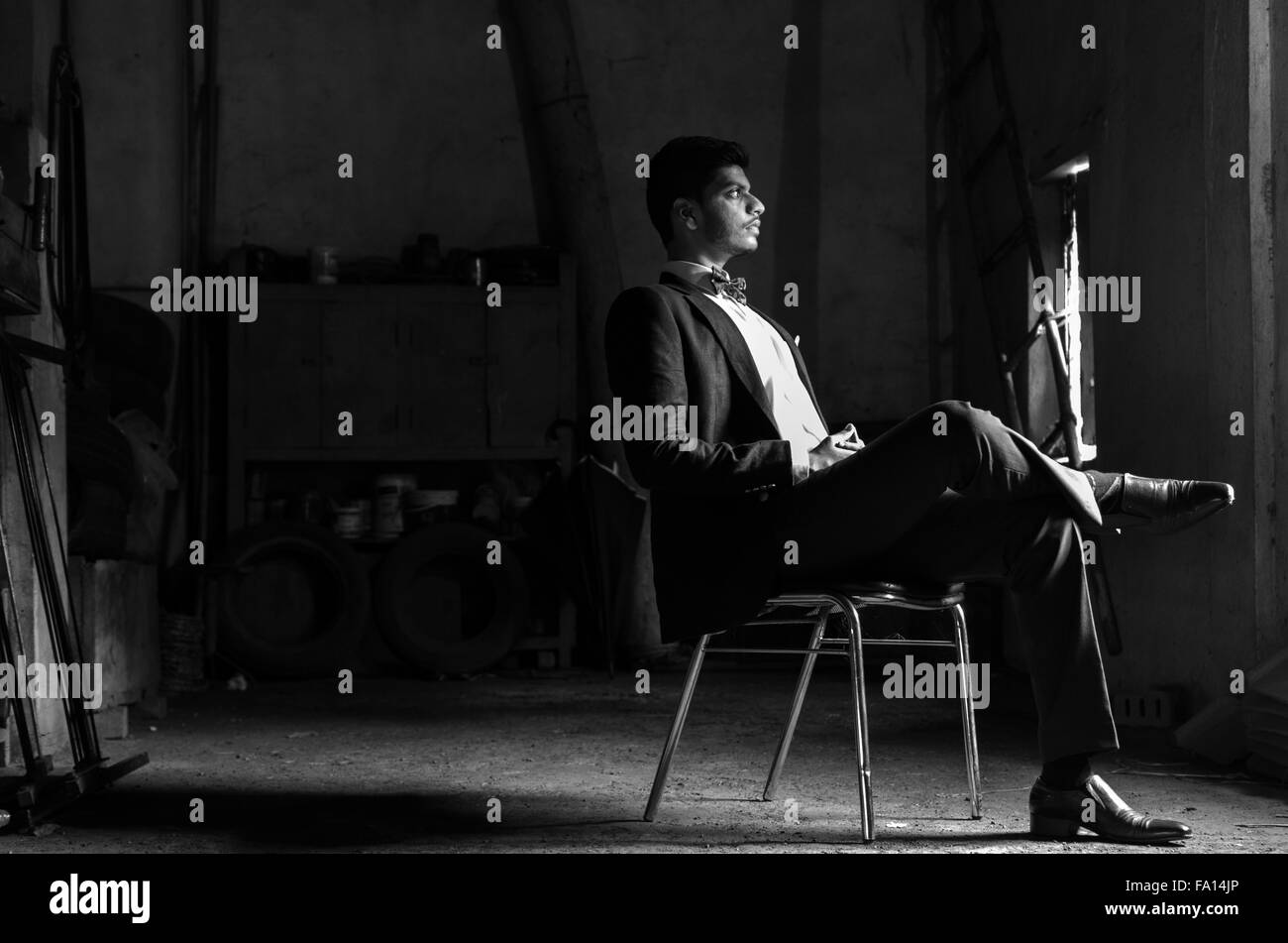 Black and White portrait of a young man dressed in black suit, sitting on a chair, in an old dark garage room. Stock Photo