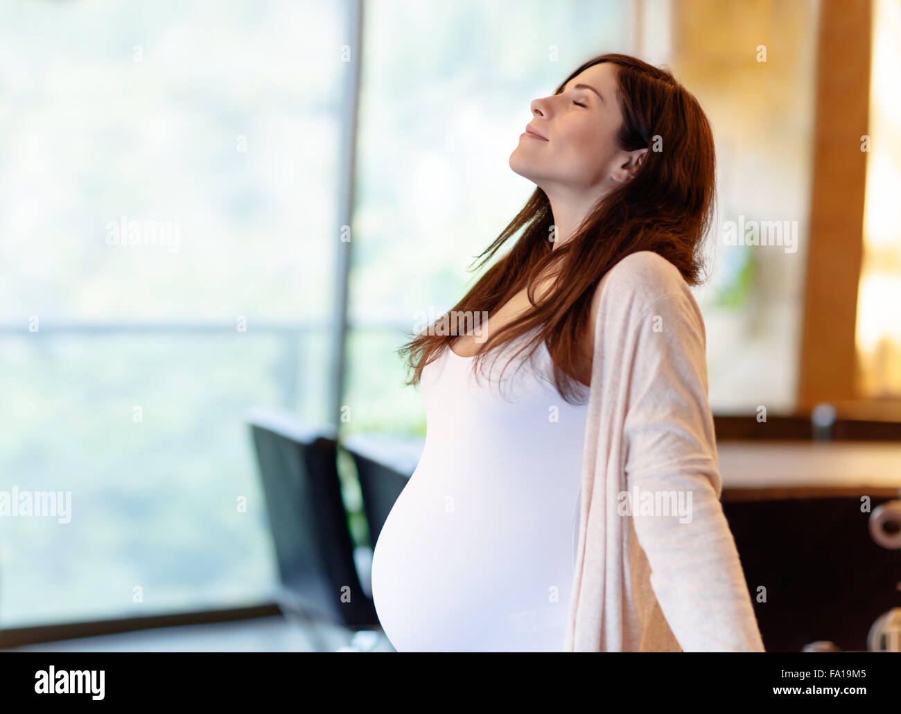 Beautiful calm pregnant woman with closed eyes standing at home in the morning and meditating, doing yoga exercise Stock Photo