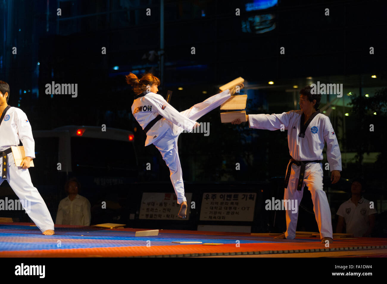A taekwondo black belt female teenager jumping in mid-air kicking and breaking a wood board held by colleague at free outdoor su Stock Photo