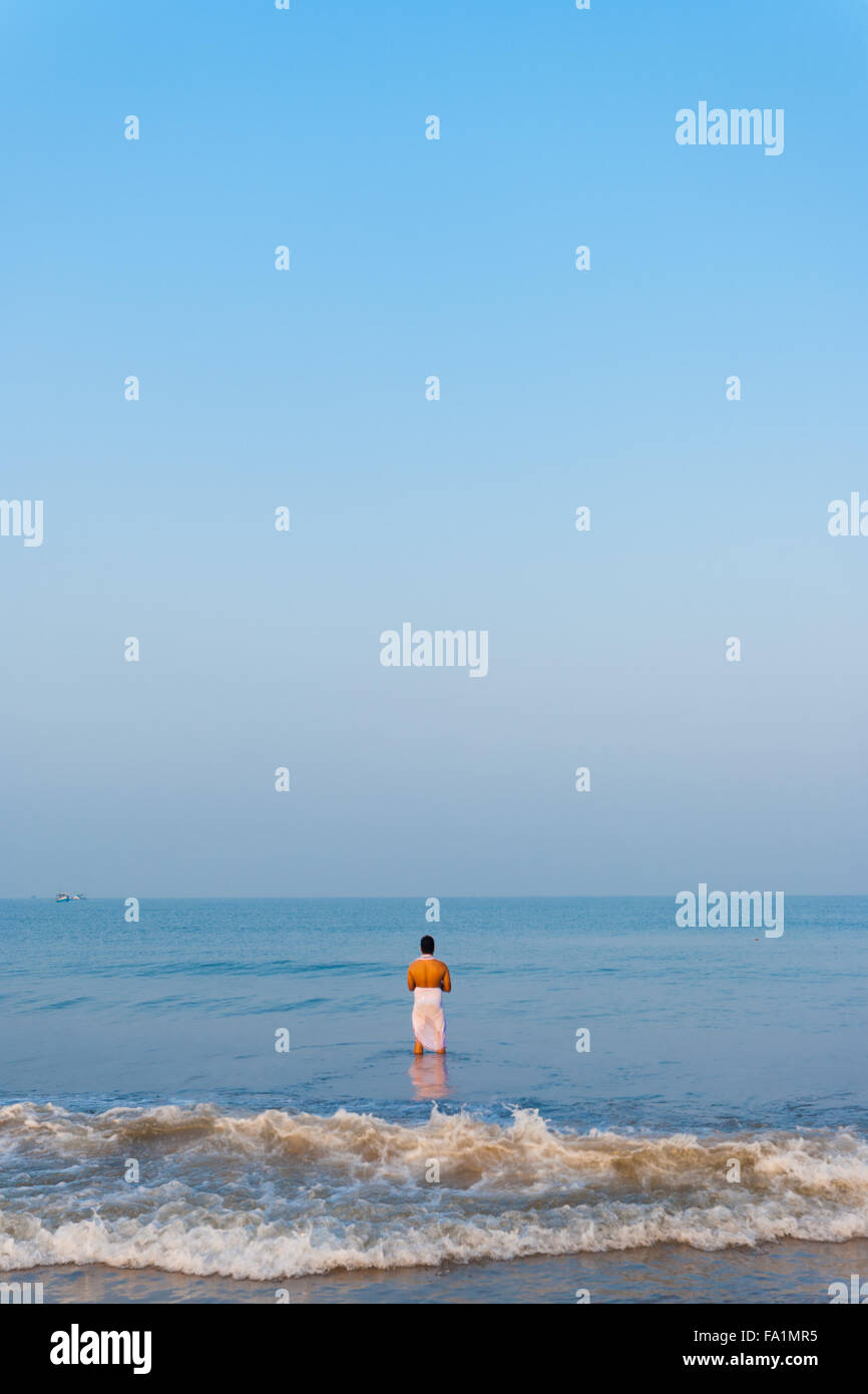 Hindu man in white lungy standing in the ocean, praying, offering gifts to Hindu gods in morning at Gokarna beach, a holy site Stock Photo