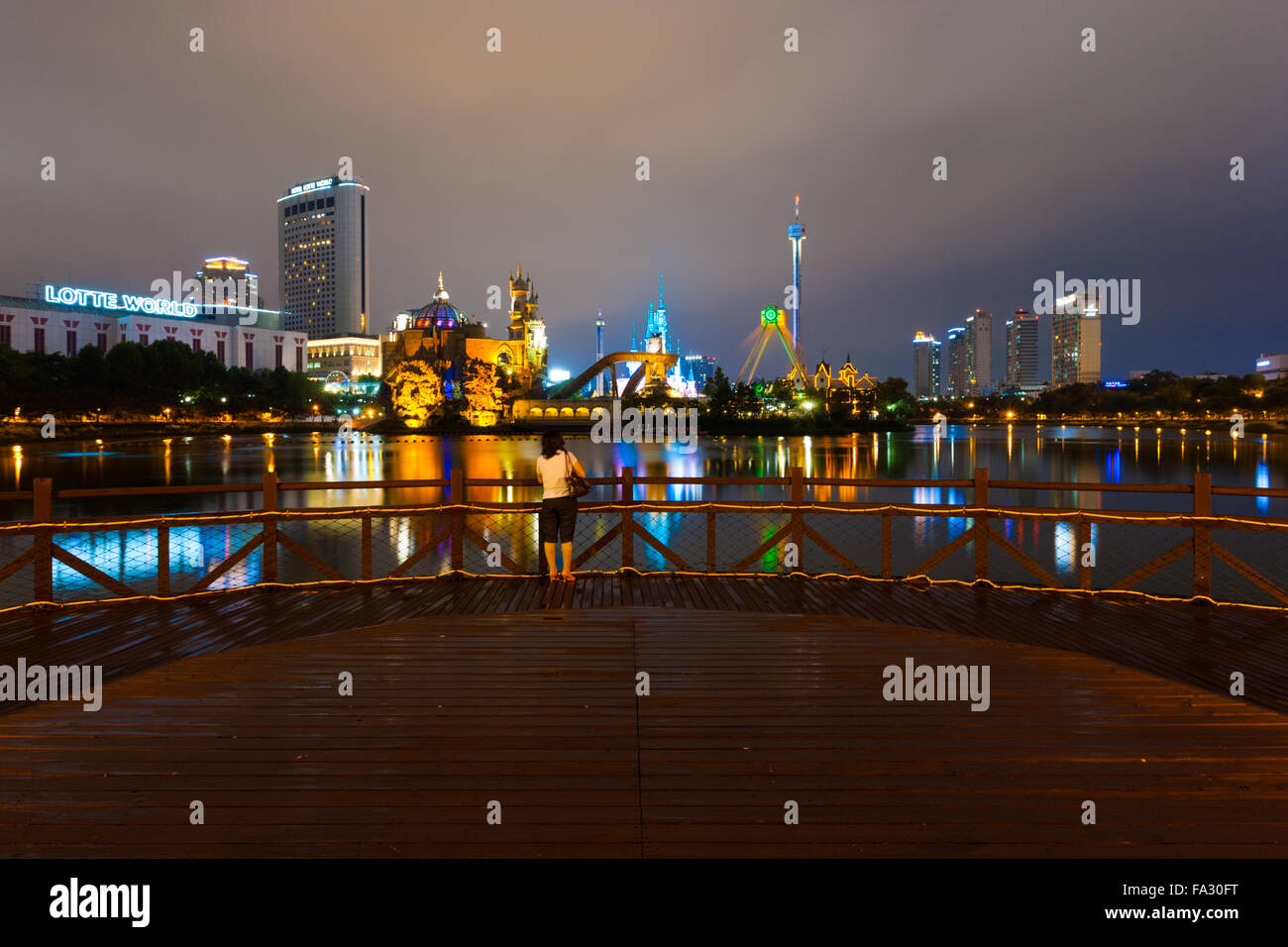 Korean woman standing on a wooden deck looking at the night lights lake water reflection at Lotte World amusement park in Seoul, Stock Photo