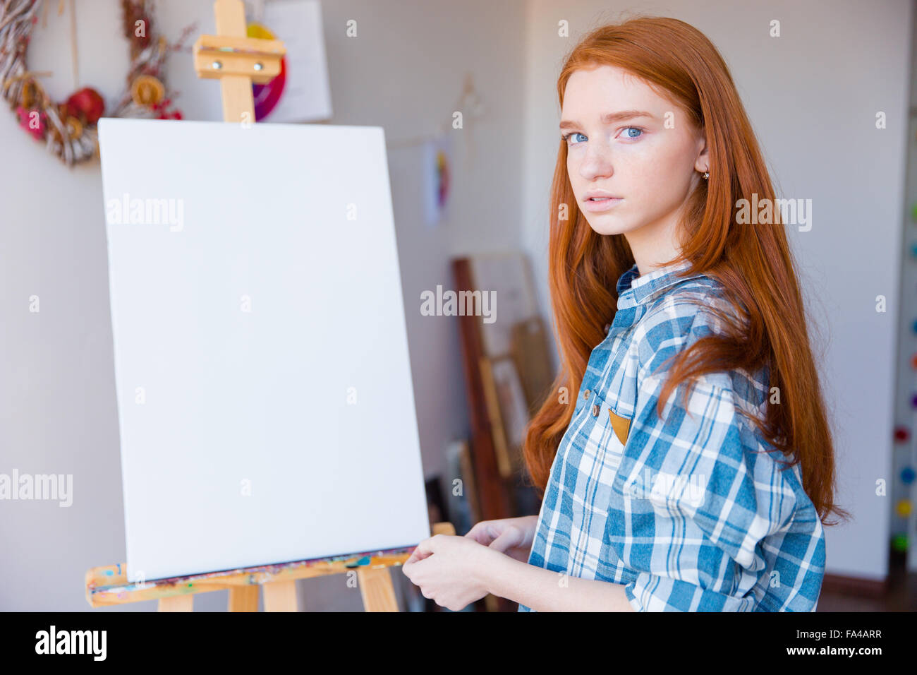 Portrait of beautiful pensive young woman painter in checkered shirt standing near blank easel in art classroom Stock Photo