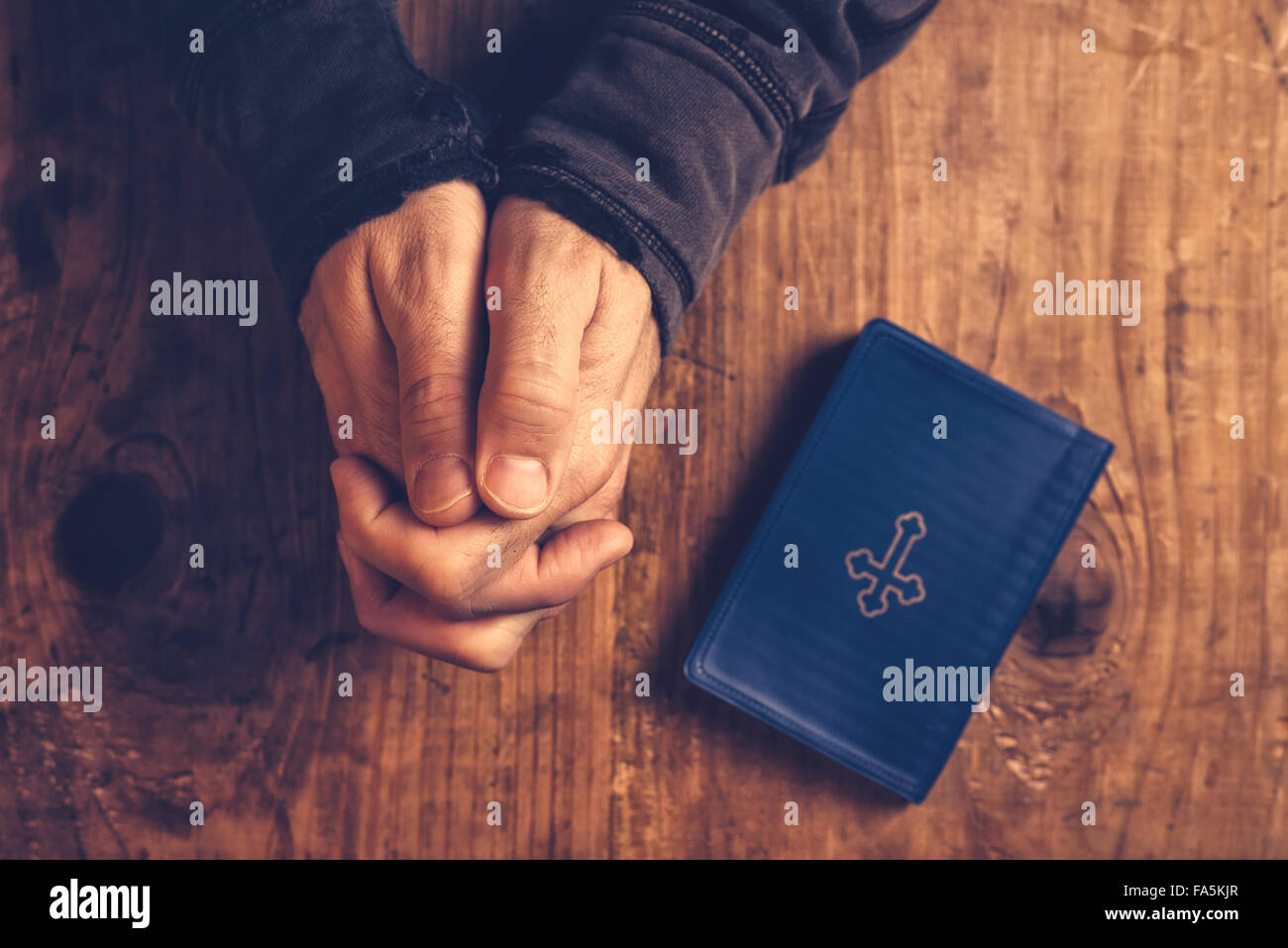 Christian man praying with hands folded and fingers crossed with Holy Bible by his side on wooden desk in church, top view Stock Photo