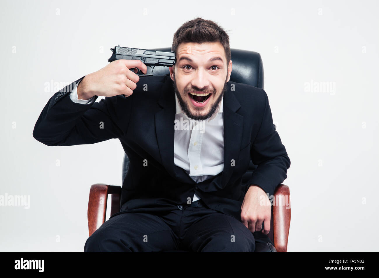Crazy hysterical bearded young businessman sitting on black office chair and put gun to his temple over white background Stock Photo