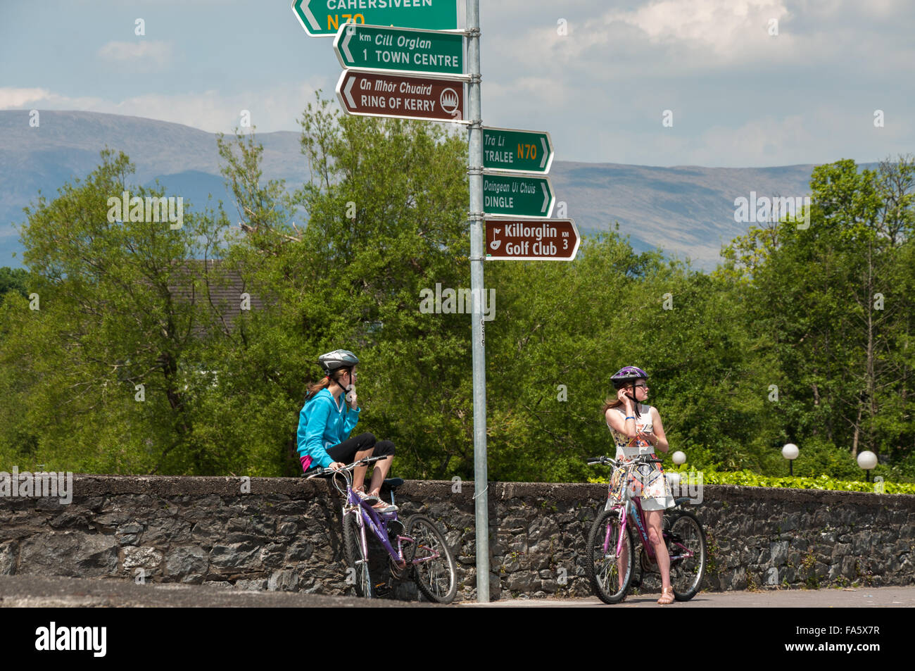 Two young women with helmets and bicycles wondering about the direction at crossroad. Road direction signs in English and Irish. Cycling in Ireland. Stock Photo