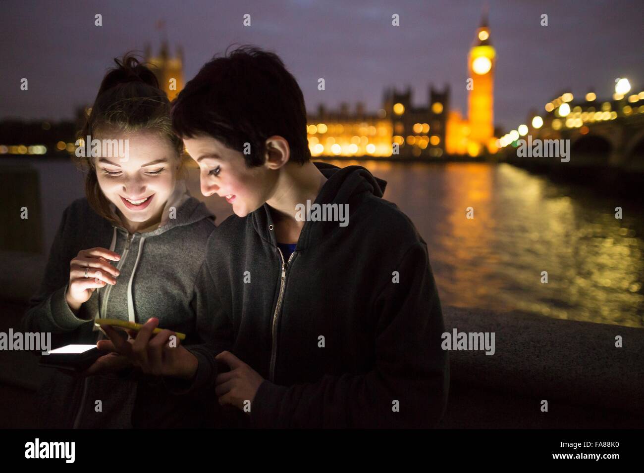 Young women illuminated by smartphone looking down smiling opposite Palace of Westminster, London, UK Stock Photo