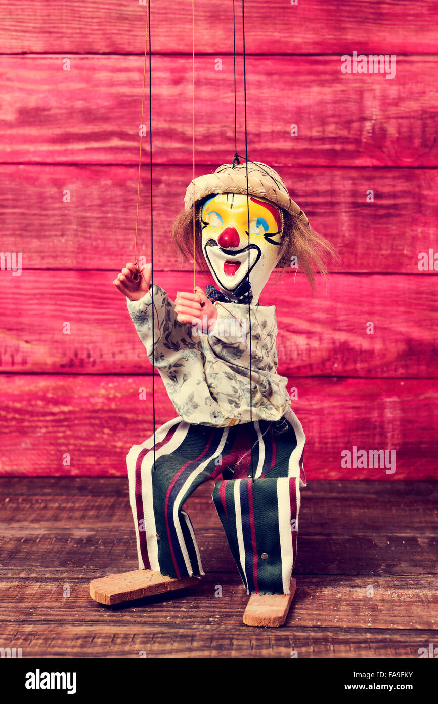 an old marionette with its face painted like a clown being manipulated on a red rustic wooden surface Stock Photo