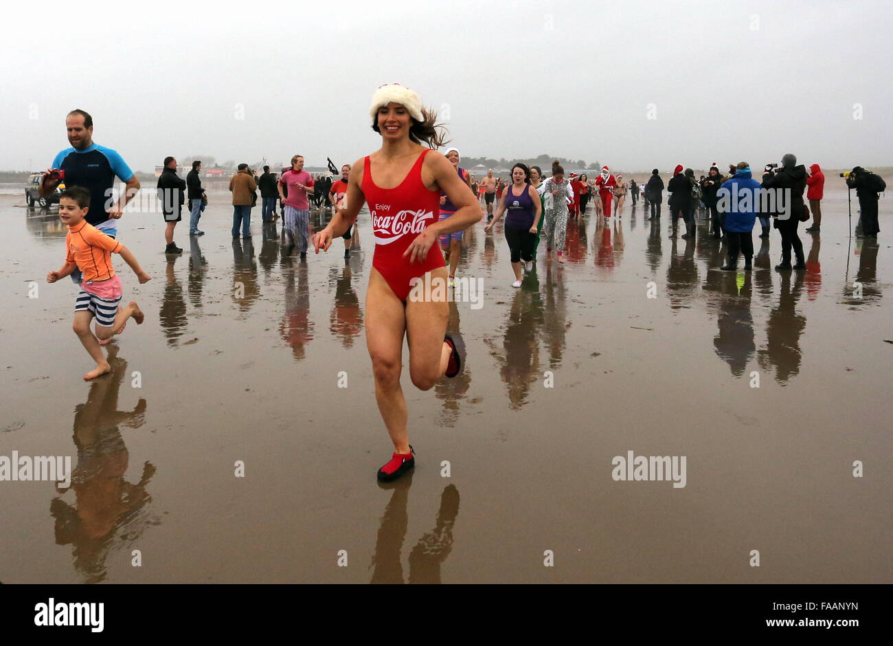 Porthcawl, UK. Friday 25 December 2015 A woman in a Coca Cola bathing costume and a Santa hat joins othert Christmas swimmers running towards the seaRe: Hundreds of people in fancy dress, have taken part in this year's Christmas Swim in Porthcawl, south Wales. Credit:  D Legakis/Alamy Live News Stock Photo