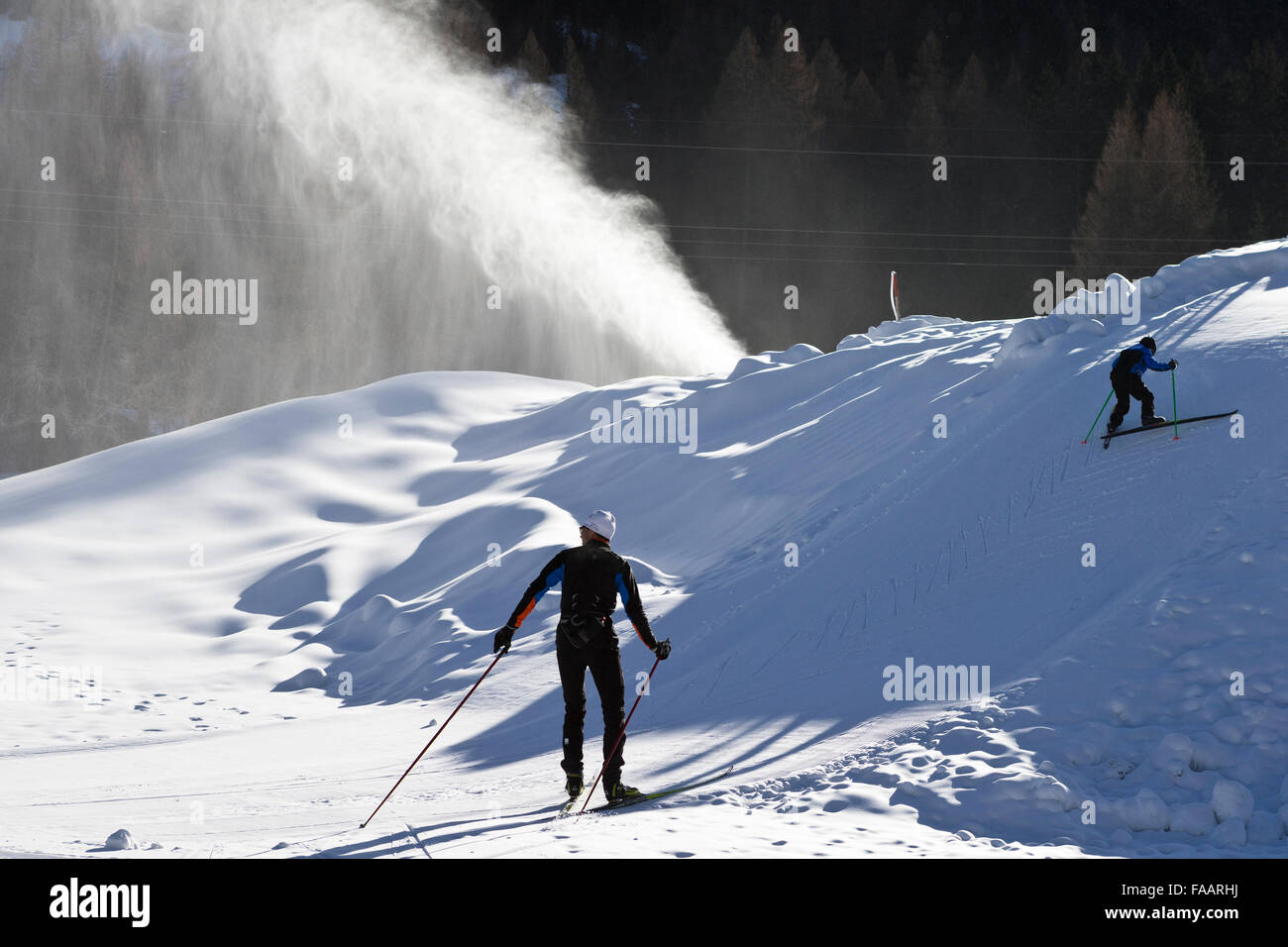 Ulrichen, Switzerland. 25th December, 2015. A snow cannon is producing artificial snow. In the foreground two crosscountry skier. There is a lack of snow in the skiing areas of Switzerland. Slopes are created with artificial snow to attract some tourists Credit:  Dominic Steinmann/Alamy Live News Stock Photo
