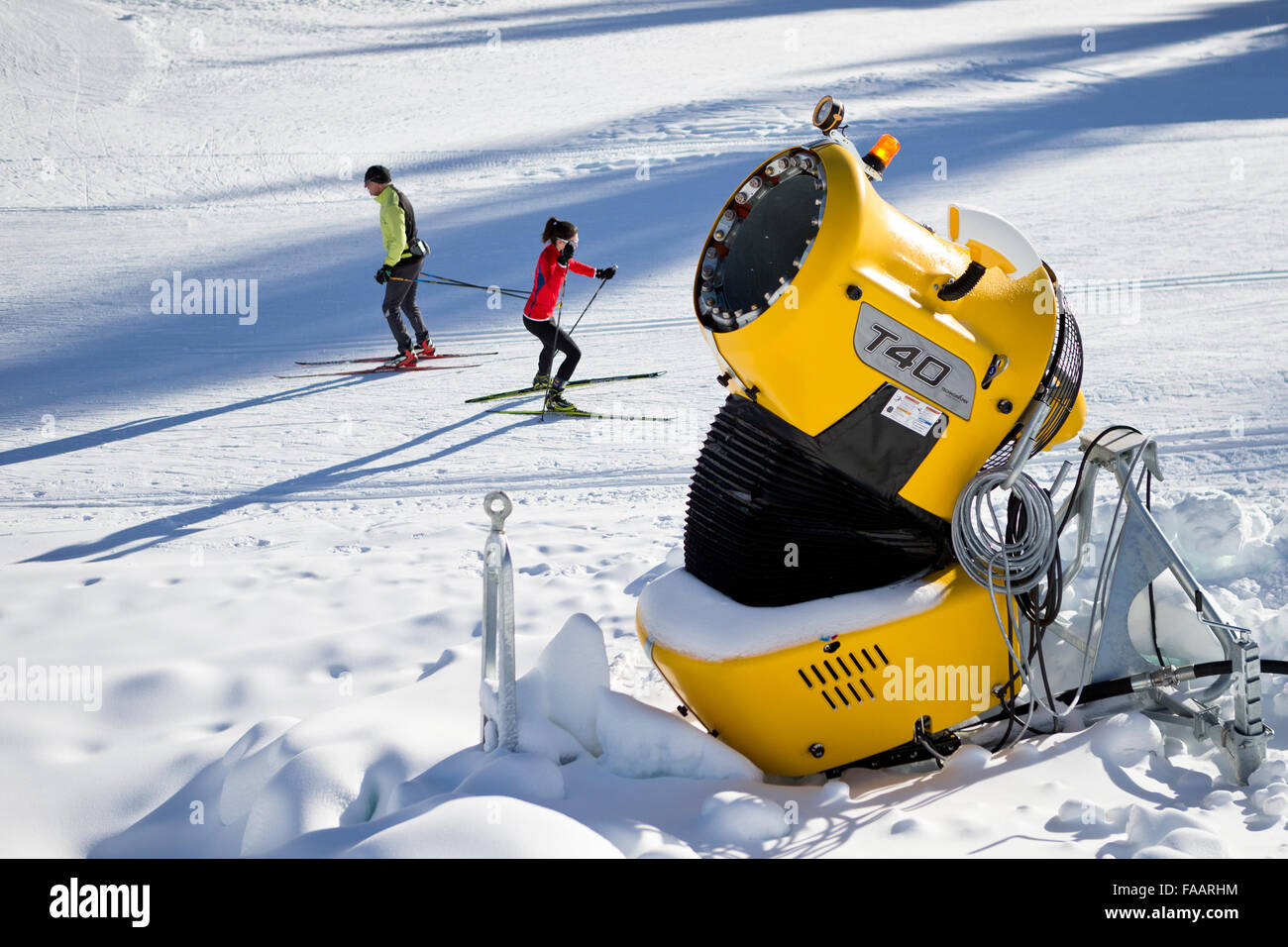 Ulrichen, Switzerland. 25th December, 2015. A snow cannon to produce artificial snow. In the background two crosscountry skier. There is a lack of snow in the skiing areas of Switzerland. Slopes are created with artificial snow to attract some tourists and reduce the economic loss. Credit:  Dominic Steinmann/Alamy Live News Stock Photo
