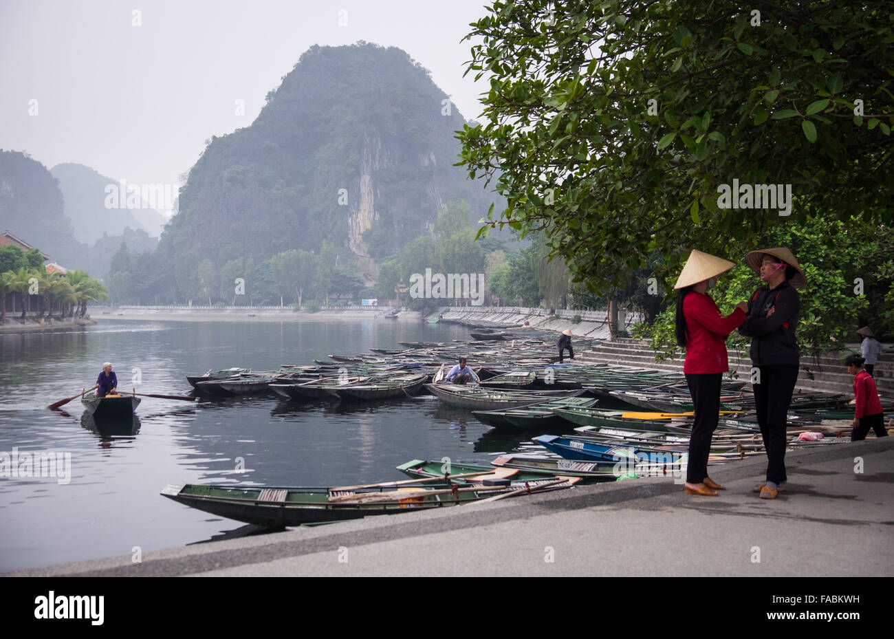 Two women wearing conical hats chatting on the quayside at Tam Coc. Approximately 1300 people work at this location, and share the job of rowing  visitors along the 5km boat ride up the river surrounded by spectacular limestone karst scenery. Stock Photo