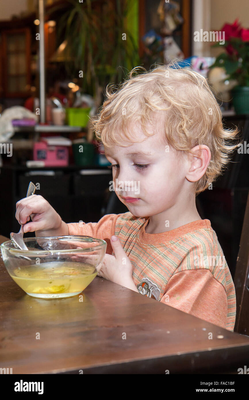A young , blond hair boy is eating soup on his own . Stock Photo