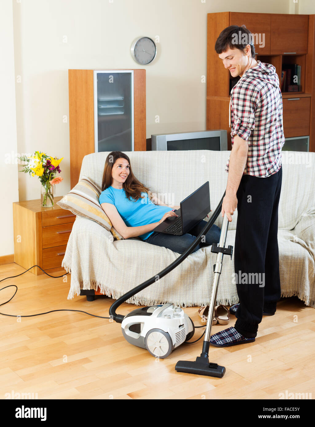 Man cleaning with vacuum cleaner  floor while woman lying with notebook over sofa at home Stock Photo