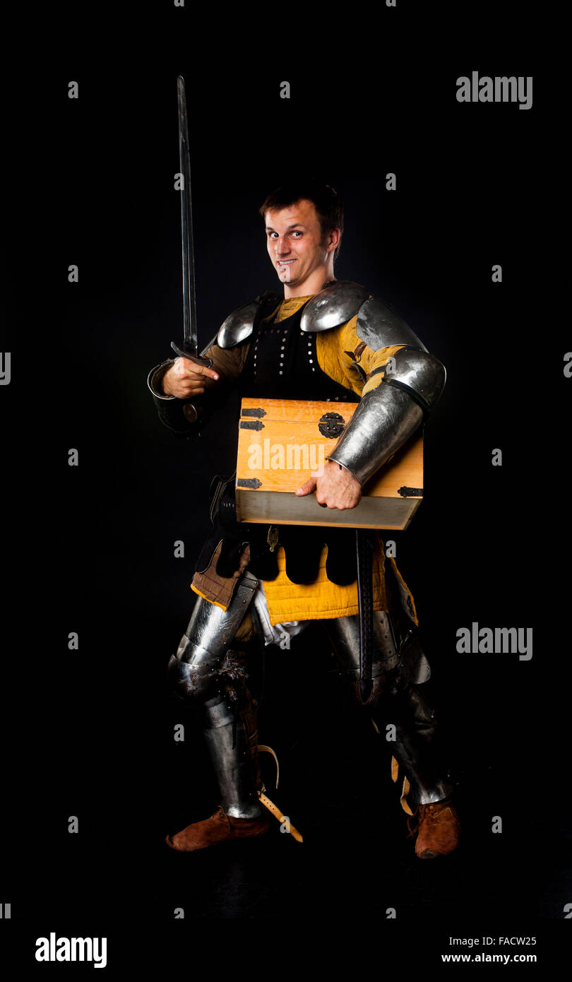 Studio shot of young man dressed as medieval knight with a sword carrying a treasure chest Stock Photo