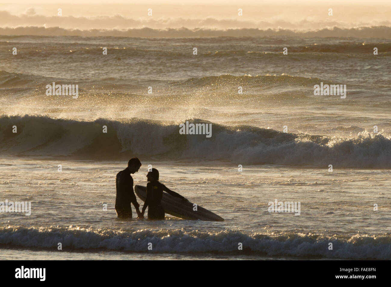 Surfer couple at sunset with waves breaking behind them. Stock Photo