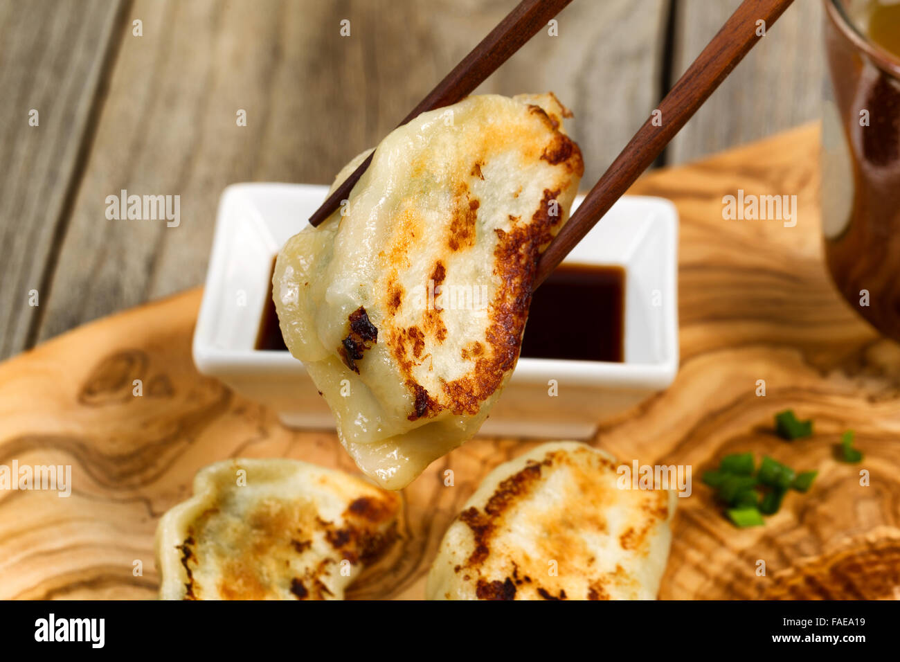 Close up view of chopsticks pickup up single cooked Chinese dumpling with soy sauce and tea in background. Stock Photo