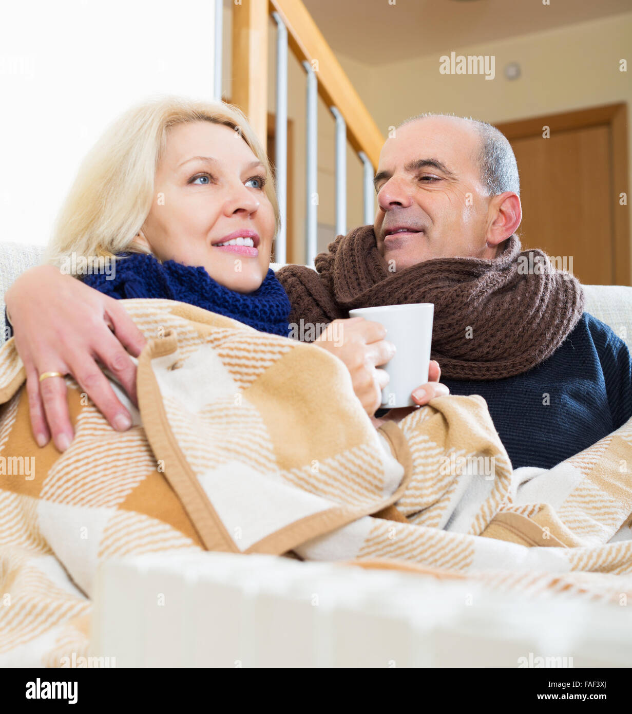 Portrait of mature frozen pensioners sitting on sofa and holding mugs Stock Photo