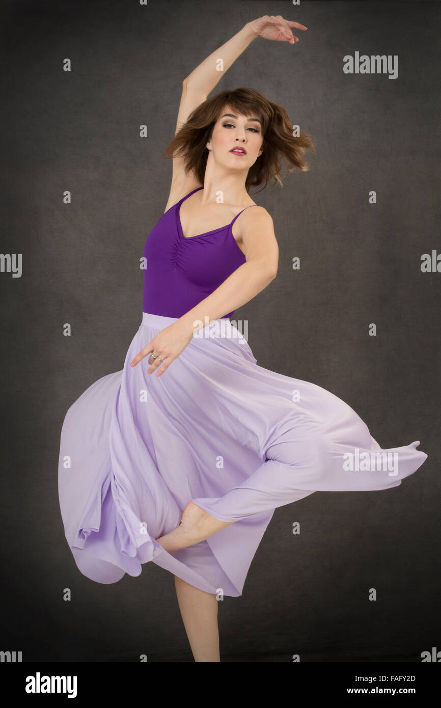 Attractive young woman dancer in purple leotard and lavender dress, pirouette turn with dress swirling, on gray background. Stock Photo