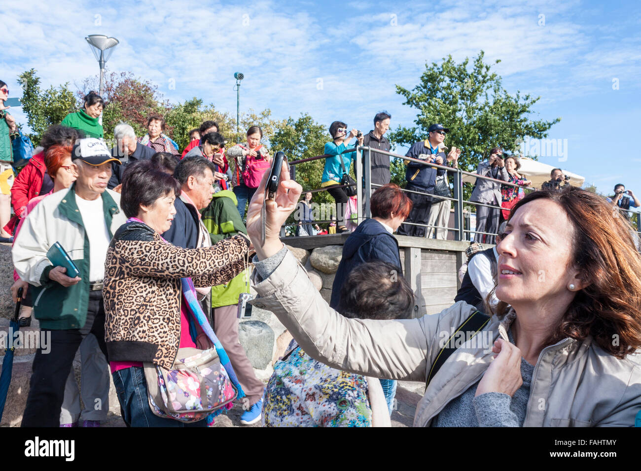 Caucasian woman surrounded by Chinese tourists takes selfie with mobile phone in front of the Little Mermaid statue, Copenhagen. Stock Photo