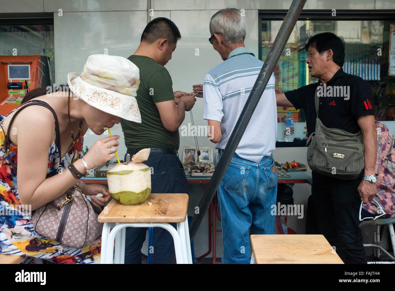 Woman drinking in a coconut in front of a Amulets stall in the street in Chinatown Bangkok, Thailand. Yaowarat, Bangkok’s Chinatown, is the World’s most renowned street food destination and the local favorite dining district. Stock Photo