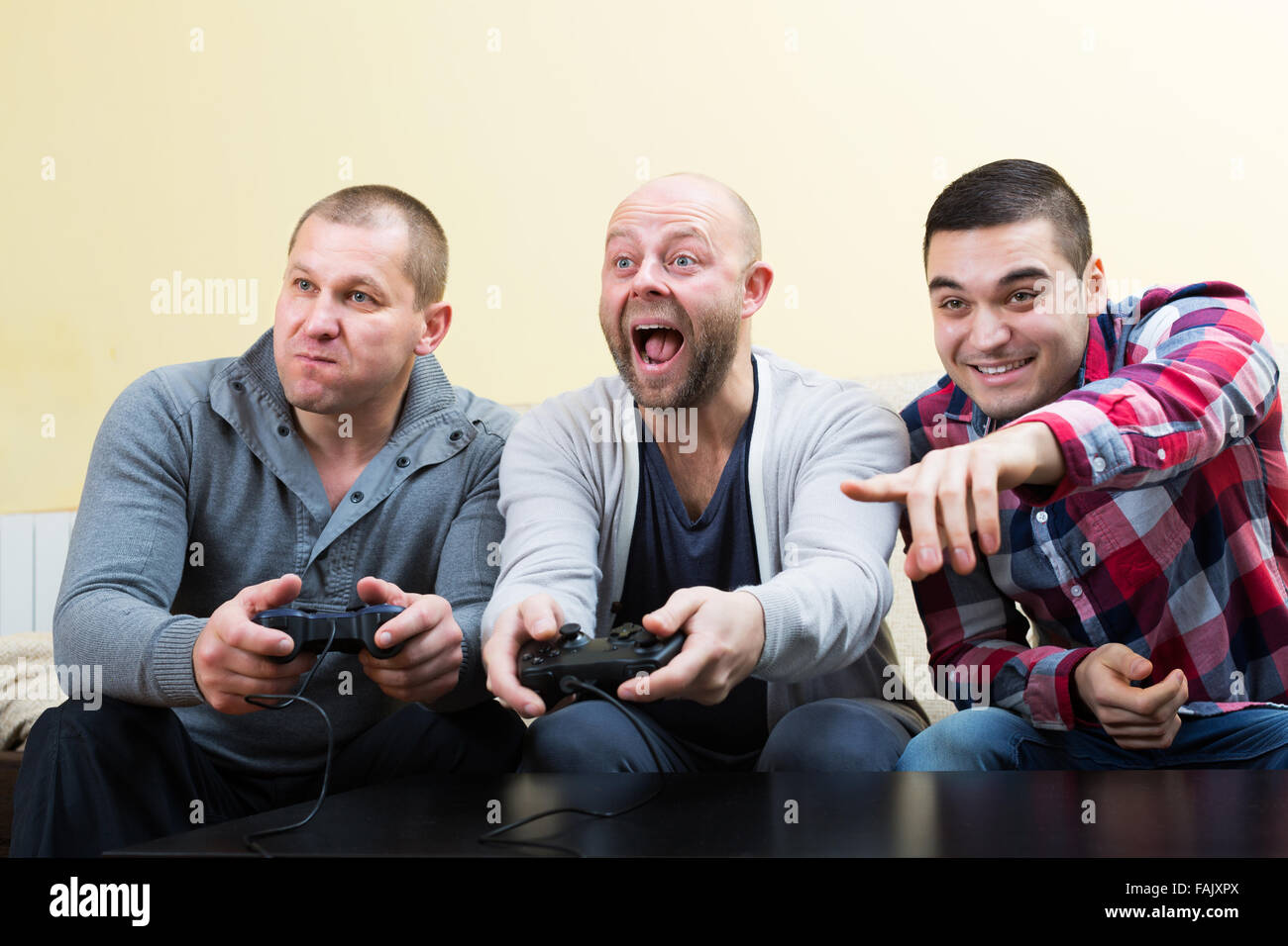 Three heated adult guys sitting on sofa with joysticks indoors Stock Photo